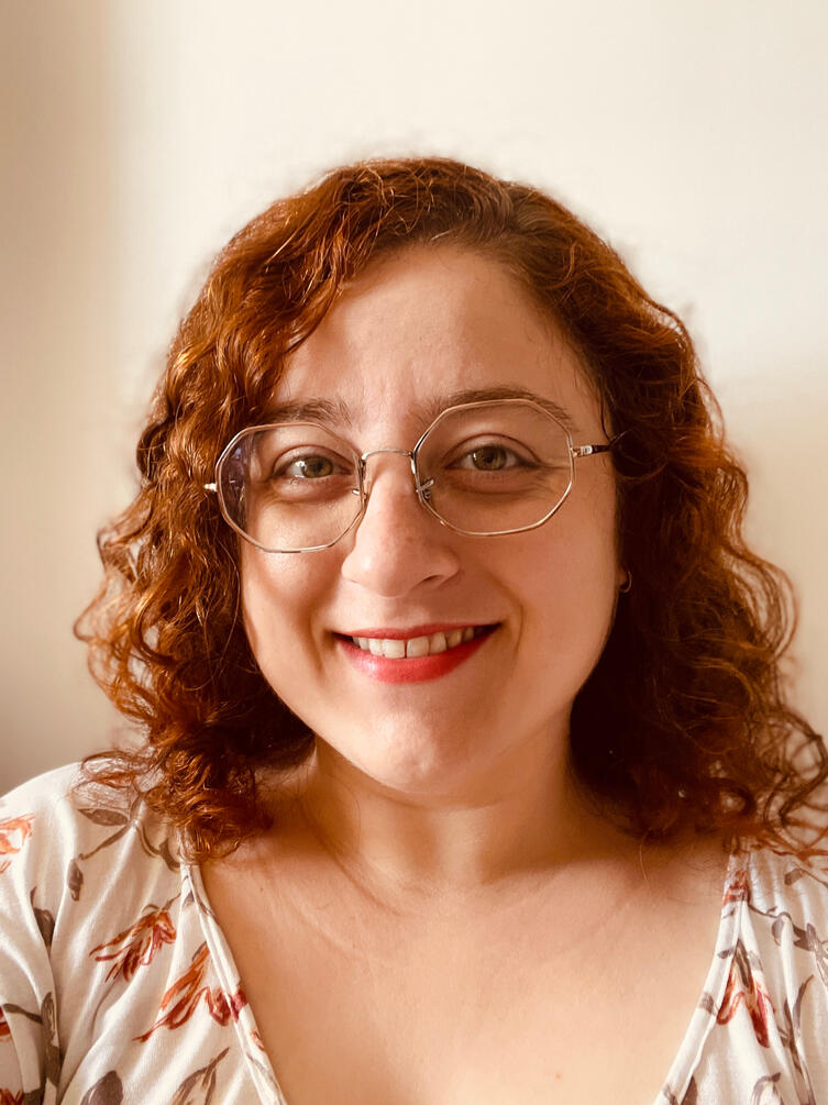 Foto de uma mulher jovem caucasiana de cabelo ondulado comprido e óculos. Ela está olhando para a câmera e sorrindo